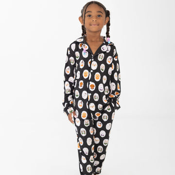 A young girl with braided hair stands barefoot against a plain white background, wearing long-sleeve two piece pajamas with dogs dressed up in costumes. 