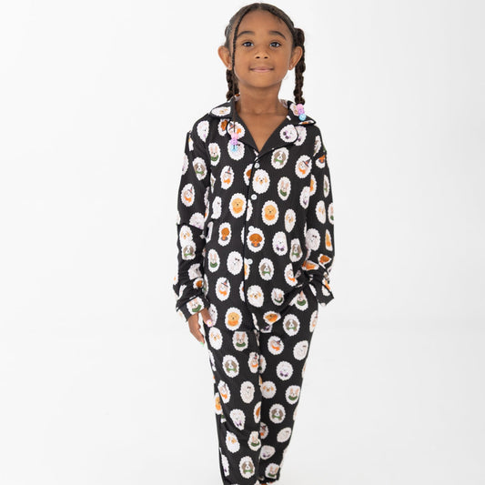 A young girl with braided hair stands barefoot against a plain white background, wearing long-sleeve two piece pajamas with dogs dressed up in costumes. 
