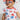 The image shows a young girl with short, curly hair standing in a bright, minimal space. She is wearing playful star-shaped glasses in red, white, and blue. Her outfit is a white T-shirt-style pajama set with flutter sleeves, decorated with a pattern of red, white, and blue bows. She appears cheerful and expressive, and the setting is softly lit with a clean, simple background that keeps the focus on her.