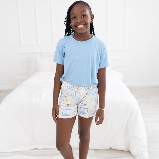 A young girl smiling in a bedroom setting, wearing a light blue short-sleeve t-shirt and the shorts have blue and white patterned featuring silly gooses in a beach theme with watermelon, beach theme and palm trees.