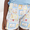 A close-up of a child wearing patterned shorts and a light blue t-shirt. The shorts feature a white base with a colorful patchwork print of cartoon ducks in swim trunks, framed like postage stamps in blue, yellow, and white.
