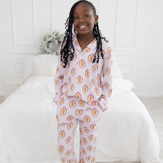 A young girl with long black braids stands barefoot in a bright bedroom, wearing a light pink striped long-sleeve pajama set with an allover gingerbread man print repeating.