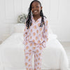A young girl with long black braids stands barefoot in a bright bedroom, wearing a light pink striped long-sleeve pajama set with an allover gingerbread man print repeating.