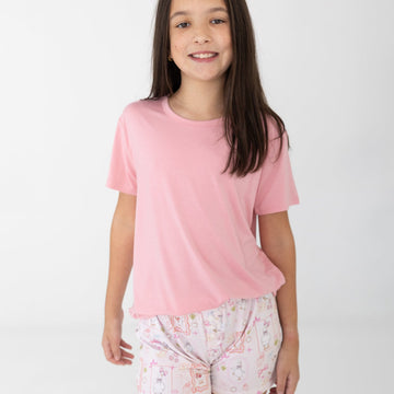 A young girl with long dark brown hair stands against a light gray studio background wearing a light pink short-sleeve t-shirt and white patterned shorts featuring silly gooses, strawberries, hearts and bows. 