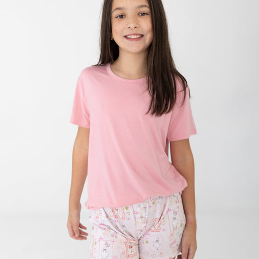 A young girl with long dark brown hair stands against a light gray studio background wearing a light pink short-sleeve t-shirt and white patterned shorts featuring silly gooses, strawberries, hearts and bows. 