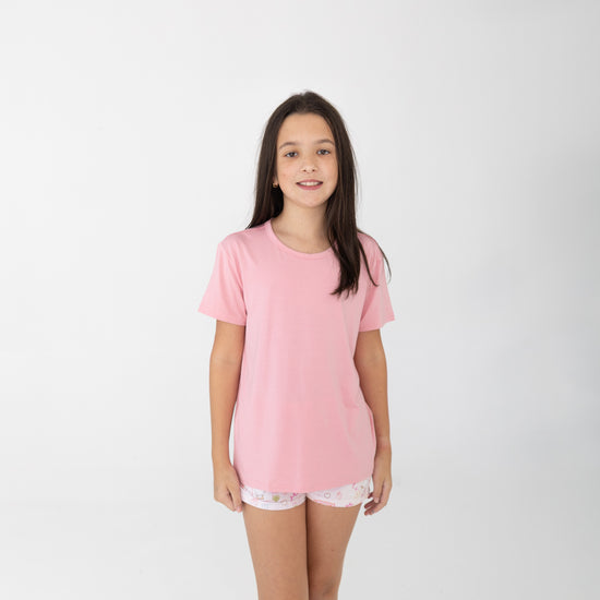 A young girl with long dark brown hair stands against a light gray studio background wearing a light pink short-sleeve t-shirt and white patterned shorts featuring silly gooses, strawberries, hearts and bows. 