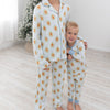 Two children standing barefoot in a bright room, wearing matching long-sleeve pajamas with a white base and a repeating gingerbread man pattern in orange and brown. A small Christmas tree is visible in the background.