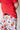 The image shows a close-up of a child wearing a bright red t-shirt and white shorts with a playful pattern. The shorts are decorated with illustrations of cows, chicken sandwiches, fries, soft drinks, and milkshakes, all in a whimsical, cartoon-like style. The child is slightly turned to the side, and their hand is resting on their hip. The background is a plain, light-colored wall.