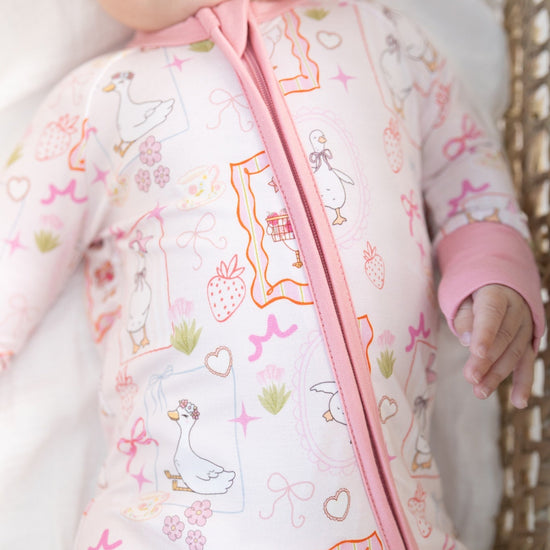 A close-up of a baby wearing a light pink one-piece sleeper with a colorful duck-themed print and a pink zipper, lying on a white blanket next to a wicker basket.