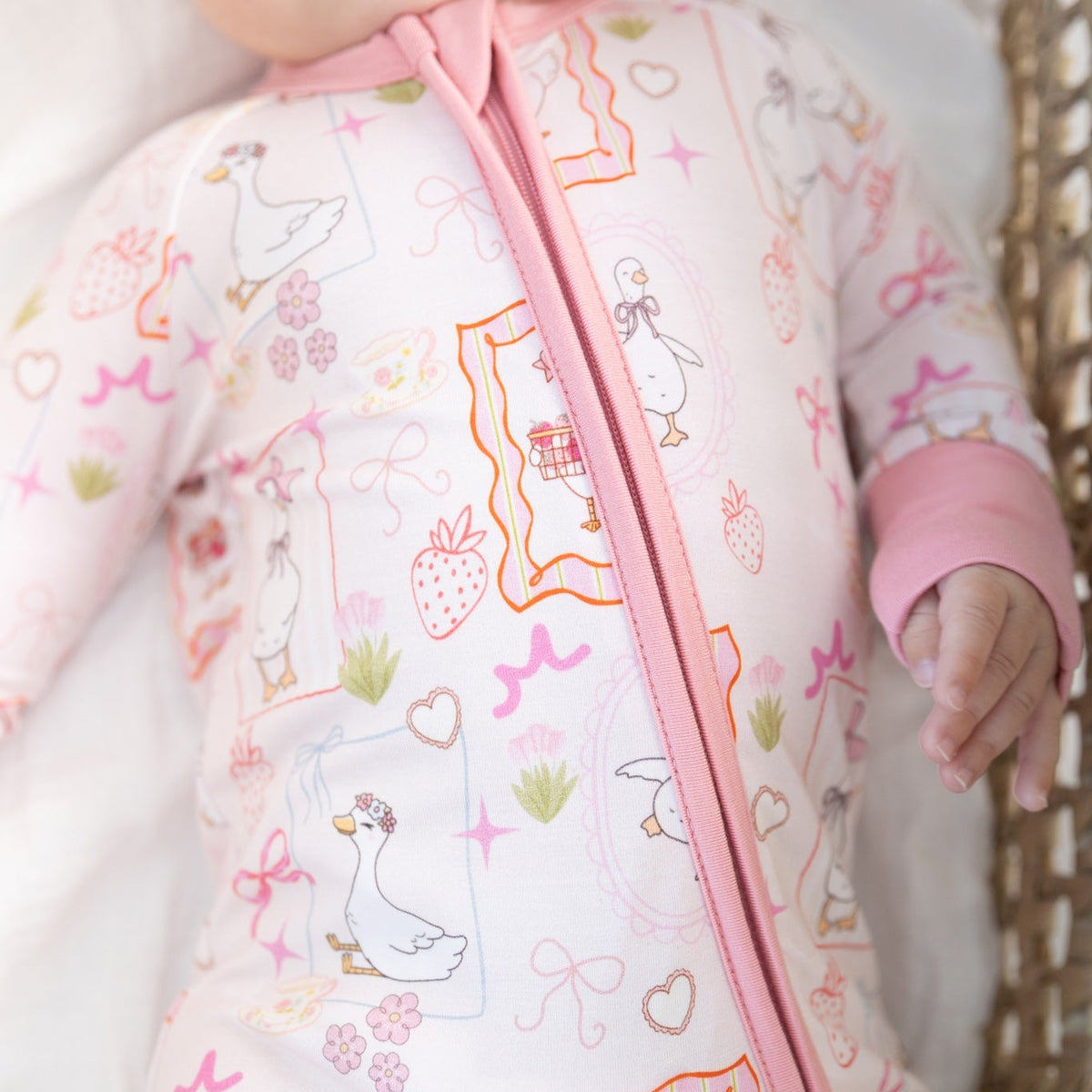 A close-up of a baby wearing a light pink one-piece sleeper with a colorful duck-themed print and a pink zipper, lying on a white blanket next to a wicker basket.