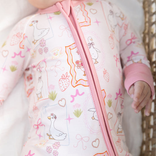 A close-up of a baby wearing a light pink one-piece sleeper with a colorful duck-themed print and a pink zipper, lying on a white blanket next to a wicker basket.