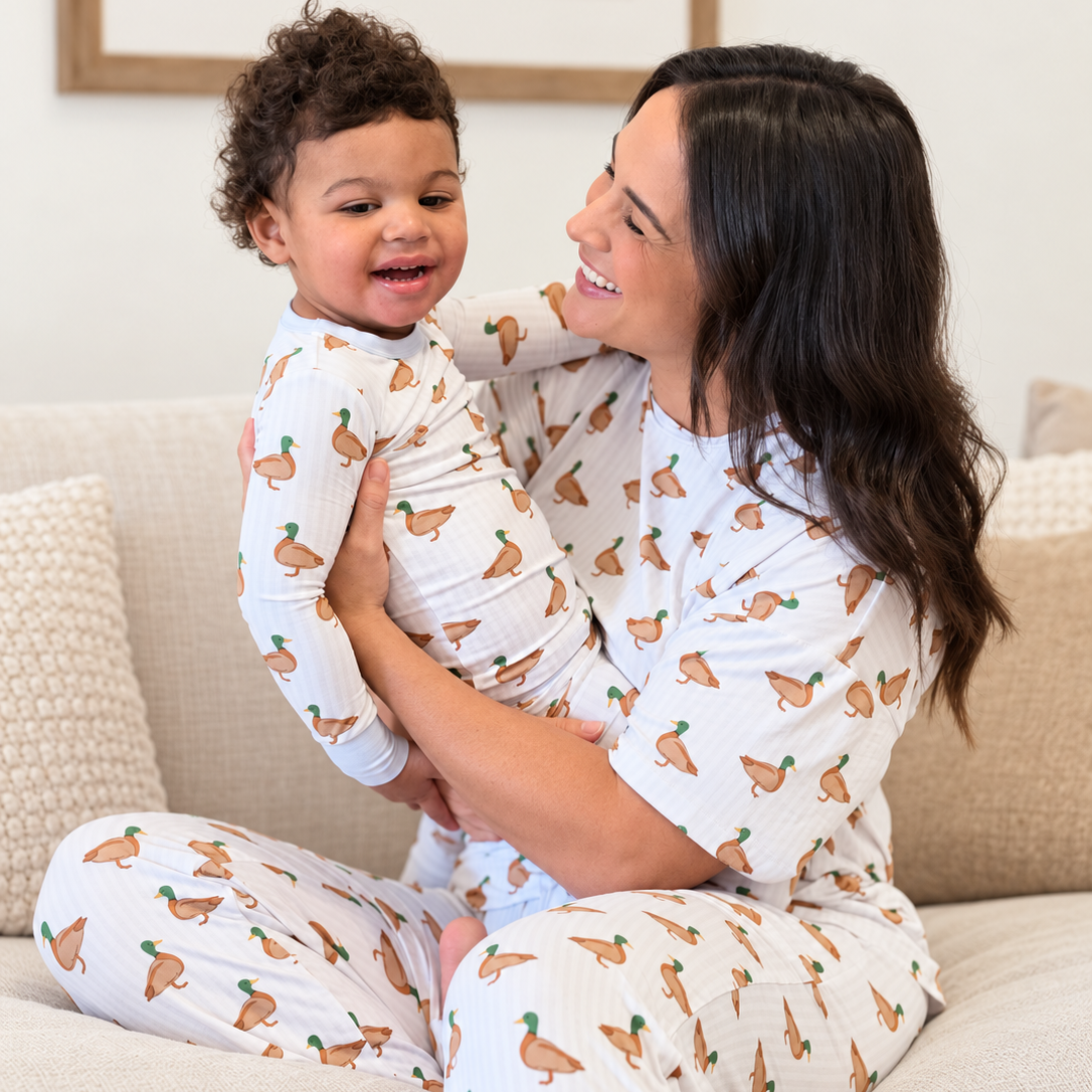 This photo shows a little boy and his mom smiling at each other and embracing on a couch. They are both wearing Monkeeroos Mallards pjs. 