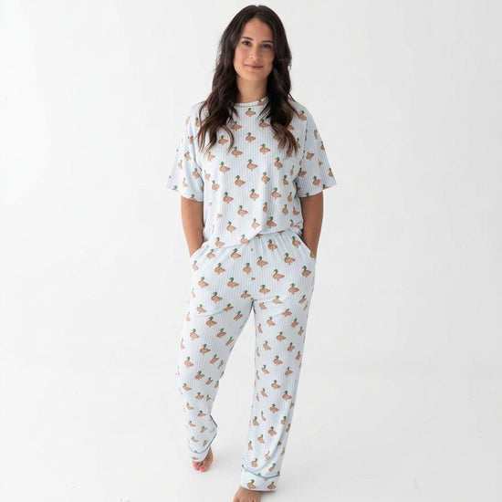 Photo shows a woman standing with her hands in her pockets wearing Monkeeroos Mallards Women's Lounge Set. Its a light background with blue trim and has mallard ducks all over it.