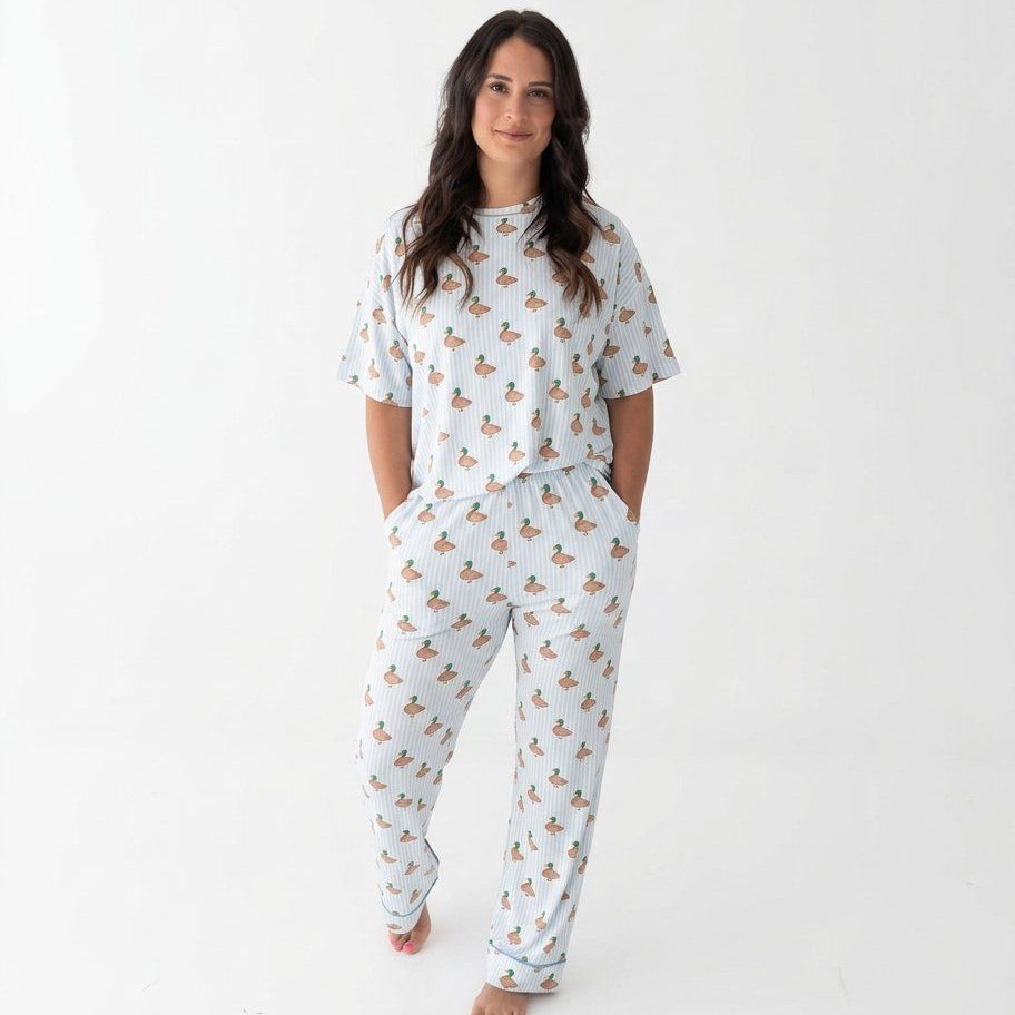 Photo shows a woman standing with her hands in her pockets wearing Monkeeroos Mallards Women's Lounge Set. Its a light background with blue trim and has mallard ducks all over it.