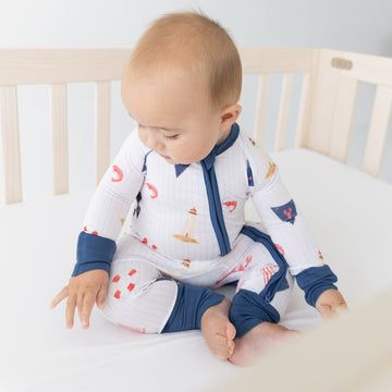 A baby sitting in a light wood crib wearing a white long-sleeve pajama with navy blue trim and a red and orange nautical print.