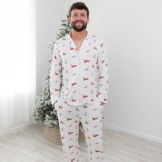 A man wearing a white long-sleeve pajama set with a red and green Christmas tree pattern, standing barefoot in a bright room with a light wood floor and a decorated Christmas tree in the background.