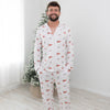 A man wearing a white long-sleeve pajama set with a red and green Christmas tree pattern, standing barefoot in a bright room with a light wood floor and a decorated Christmas tree in the background.
