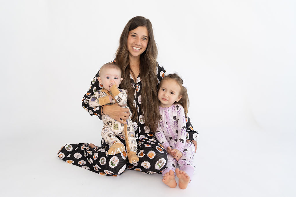 A woman and two children sitting on a white studio floor, wearing matching patterned pajamas. The woman holds a baby on her lap, and the child on the right wears a light purple pajama set.