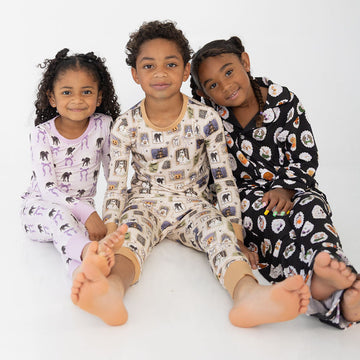 Three children sitting together on a white studio floor wearing matching long-sleeve pajamas with animal prints. The child on the left wears light lavender pajamas with black animal silhouettes, the center child wears cream pajamas with multicolored animal illustrations, and the child on the right wears black pajamas with white and orange animal illustrations.