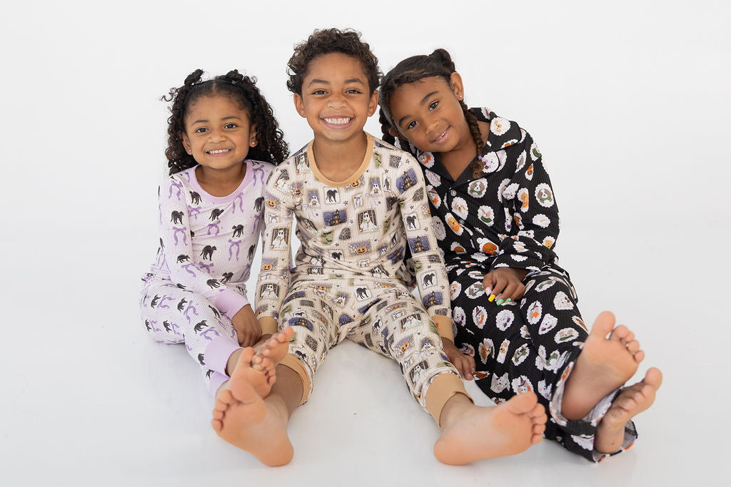 Three young girls sitting together on a white studio floor, wearing matching pajama sets with different animal prints: lavender with pink elephants, cream with multicolor animals, and black with white animals.
