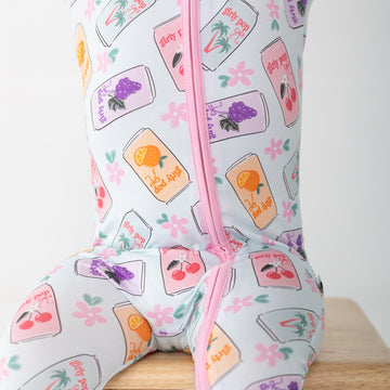 A child wearing a light gray footed zip-up sleeper with a colorful fruit and flower print, sitting on a wooden stool against a white background.