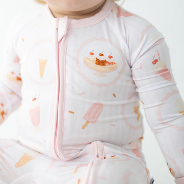 A close-up of a baby wearing a light pink long-sleeve zip-up sleeper with a playful ice cream and popsicle print in soft peach and orange tones.