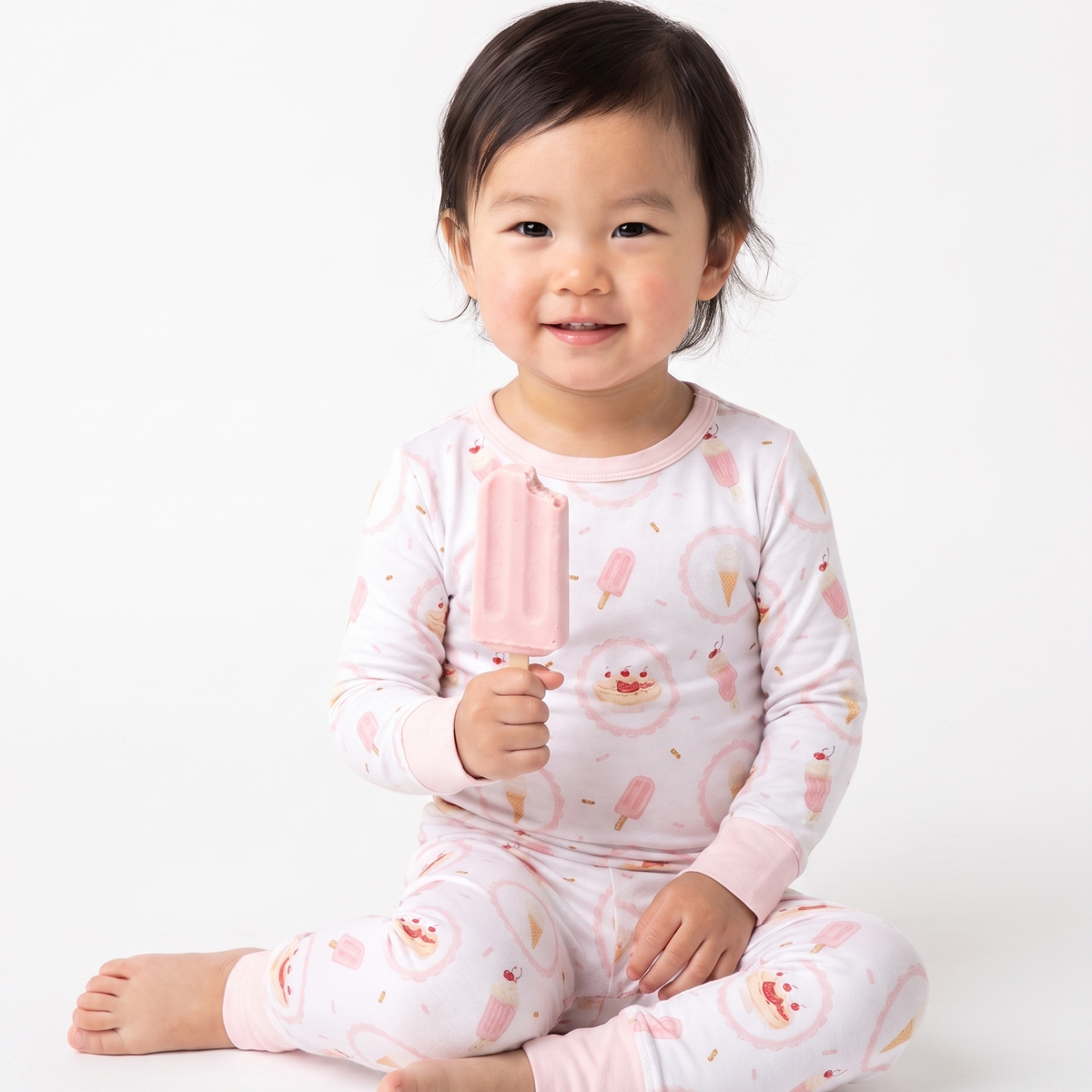 A smiling toddler girl sitting cross-legged on a white studio background, wearing a light pink long-sleeve pajama set with a playful ice cream cone and popsicle print, while holding a pink popsicle.