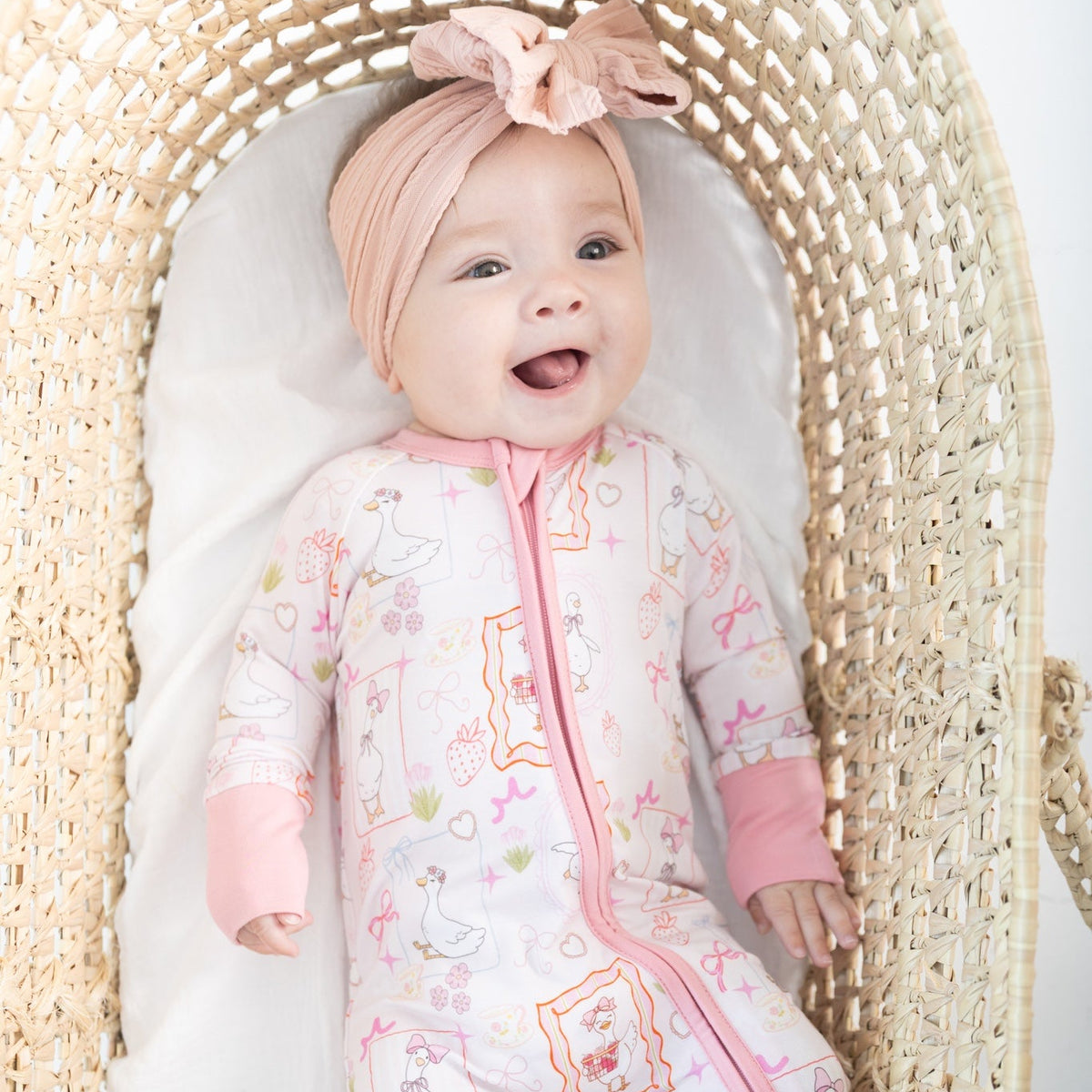 A baby lying on their back inside a woven Moses basket, wearing a pink and white patterned romper with a pink zipper featuring silly gooses, strawberries, hearts and bows.