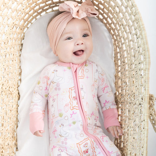 A baby lying on their back inside a woven Moses basket, wearing a pink and white patterned romper with a pink zipper featuring silly gooses, strawberries, hearts and bows.